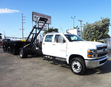 Truck side view showing a white 2023 Chevrolet Silverado 5500HD with a black roll-off dump body and hydraulic ladder/backing equipment attached.