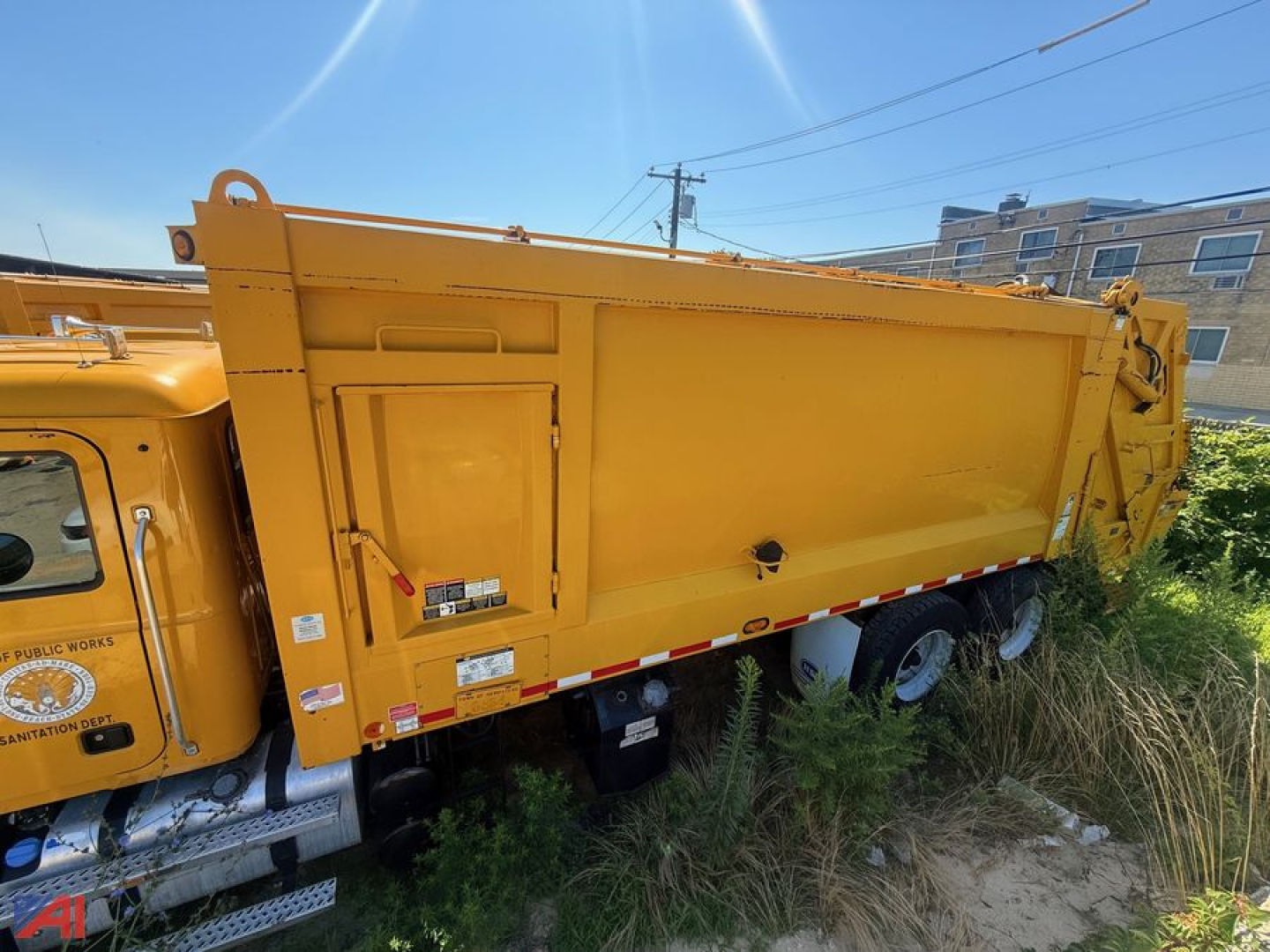 Side view of a yellow 2014 Mack garbage truck rear body with hydraulic lift, cargo container, and wheels visible; generic street scene in background.
