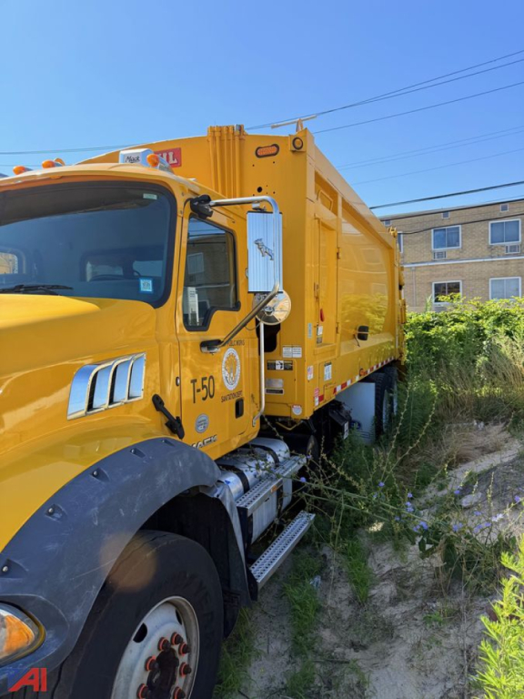 Front-left side of a yellow Mack garbage truck with T-50 label, side steps, and large rear compactor body showing equipment livery.