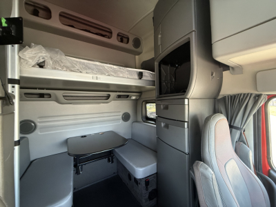 Interior sleeper cab of a 2024 Volvo VNL64T860 showing upper bunk, dinette table, storage cabinets, and passenger seat; right side has a vertical cabinet and seat with light gray upholstery.