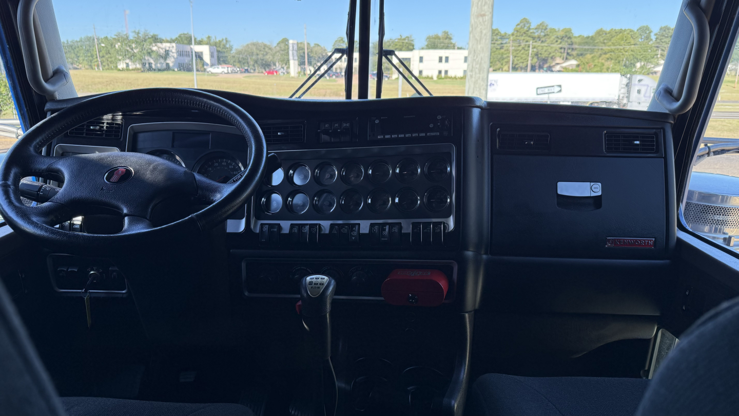 Kenworth W900 cab interior view showing steering wheel, dashboard with numerous gauges, transmission shifter, and center controls.