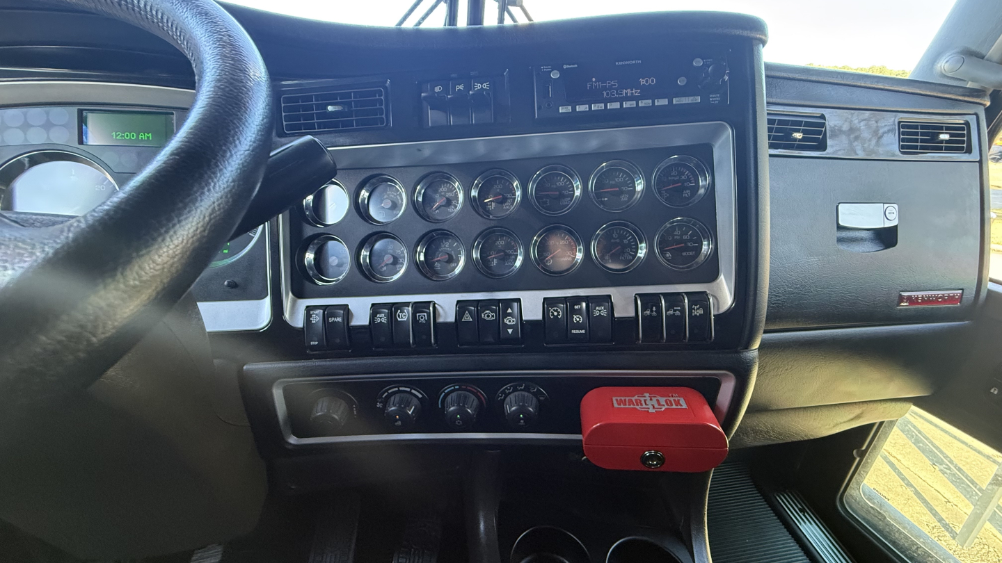 Kenworth W900 cab interior showing dashboard gauges and control switches with red device attached under dash