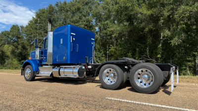 Kenworth W900 tail/side view showing blue sleeper cab and dual rear axles with chrome wheels.