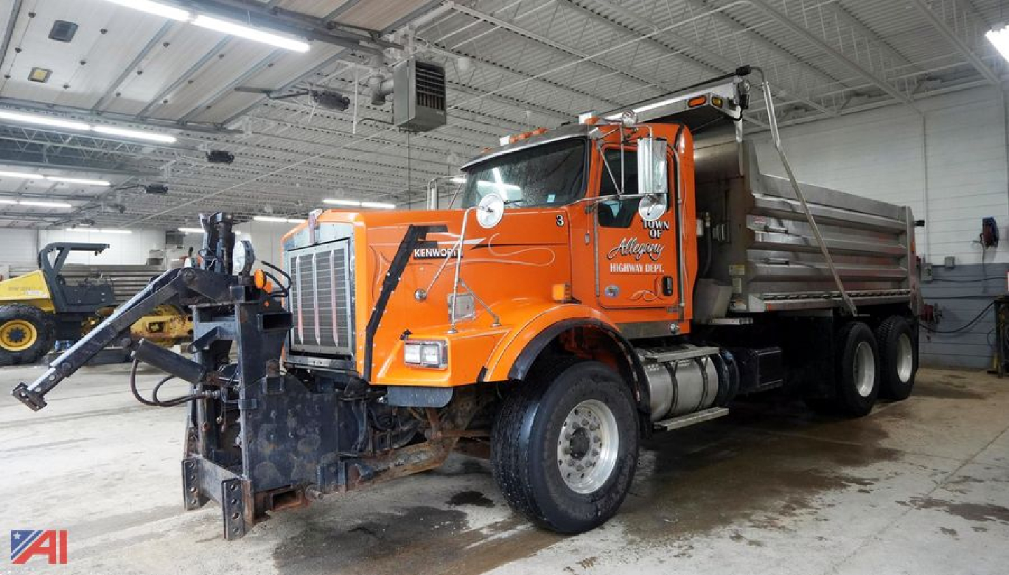 Kenworth T800 dump truck with plow attached inside a service bay, orange with dumping bed.