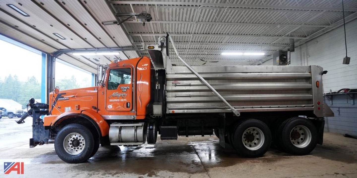 Kenworth T800 dump truck with plow attachment, orange cab and silver dump bed parked inside workshop.