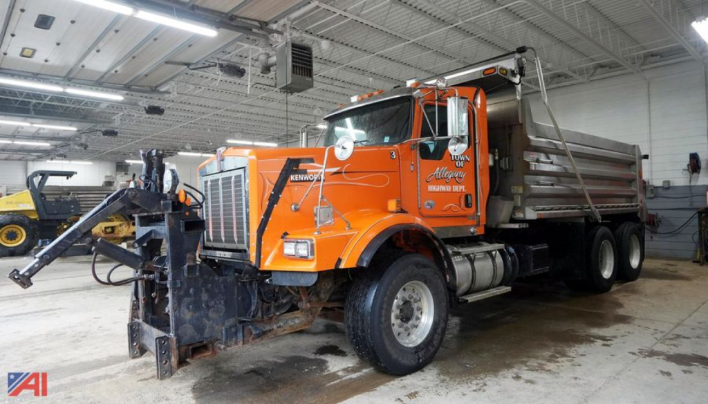 Kenworth T800 dump truck with plow attached inside a service bay, orange with dumping bed.