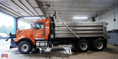 Kenworth T800 dump truck with plow attachment, orange cab and silver dump bed parked inside workshop.
