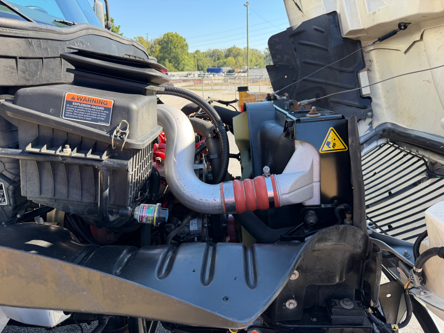 Trucks engine bay of a 2022 International MV607 showing air intake, hoses, and battery area with warning labels and intake duct.