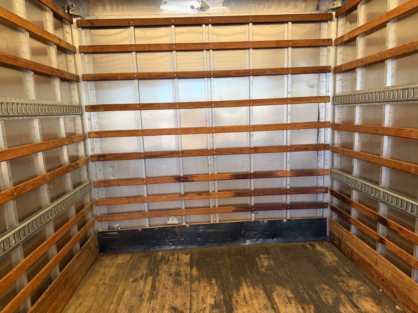 Interior of an empty box truck cargo area with wooden and metal-ribbed walls, dark floor, and overhead lighting.