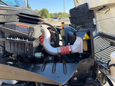 Trucks engine bay of a 2022 International MV607 showing air intake, hoses, and battery area with warning labels and intake duct.