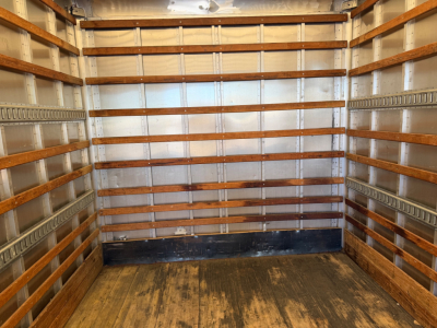 Interior of an empty box truck cargo area with wooden and metal-ribbed walls, dark floor, and overhead lighting.