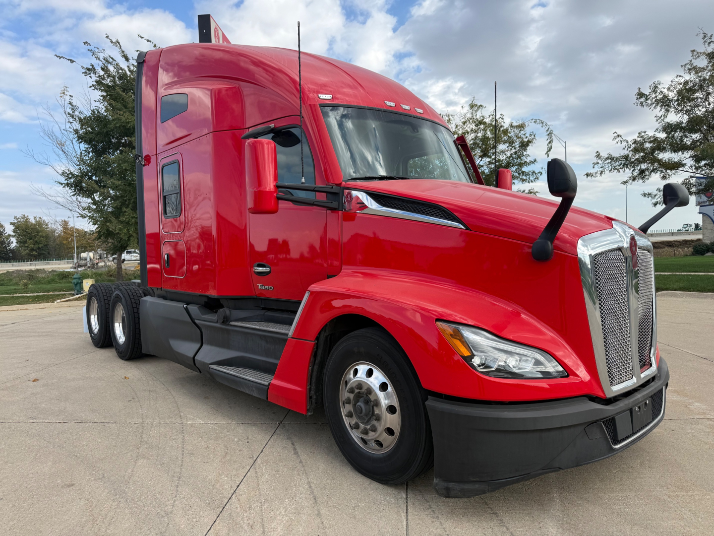 Kenworth T680 sleeper cab tractor in red, front-left view showing cab, chrome grille, large side mirrors, and dual rear axles.