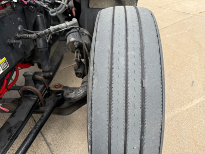 Kenworth truck engine bay with visible front tire and chassis components; large treaded front tire mounted; various hoses and wiring around.