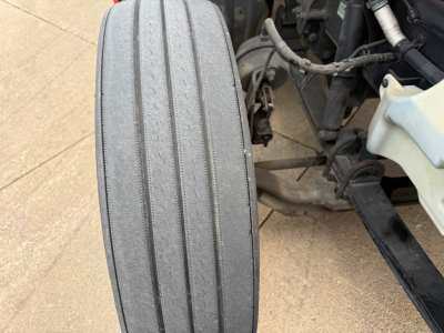 Truck undercarriage with front axle and large flat front tire viewed from side showing tire wear and suspension components.