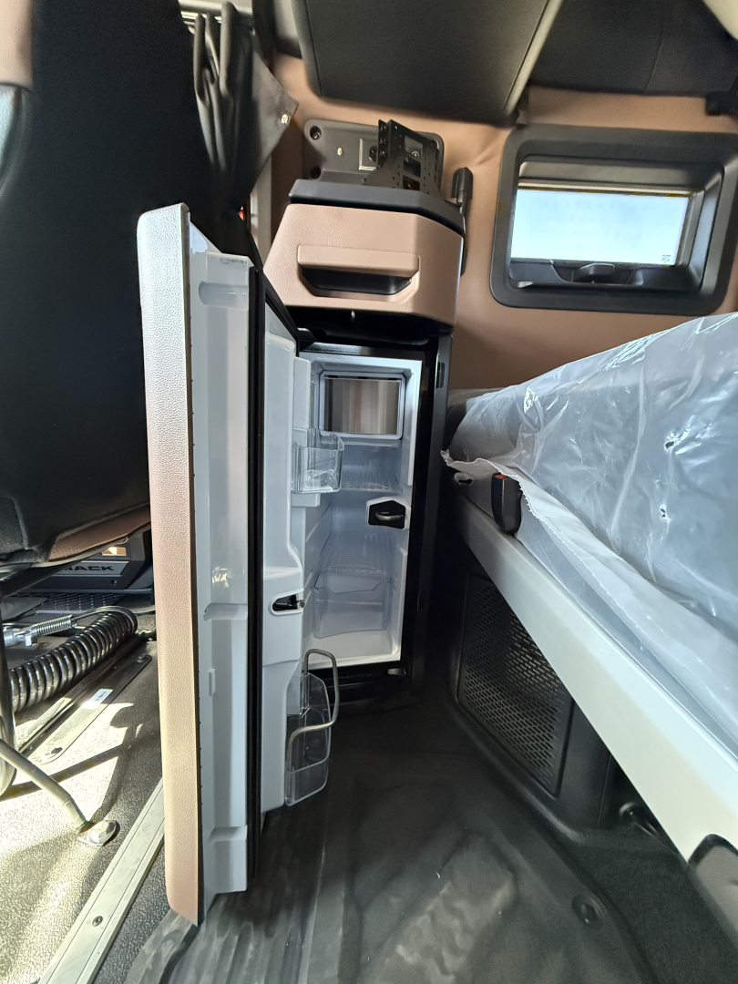Interior cabin view of a 2027 Mack AN64T64 showing a compact refrigerator/freezer unit mounted near the driver area with open doors and storage compartments.