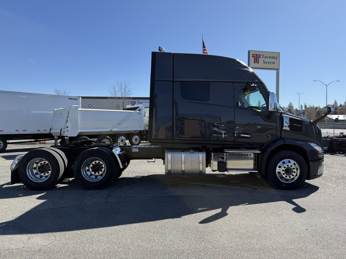 Truck side view showing a black Mack AN64T64 conventional sleeper with a white dump trailer hitched, aluminum fuel tanks, and chrome wheels.