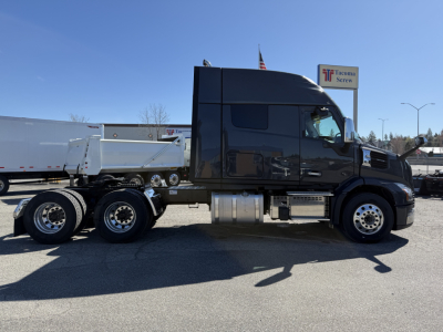 Truck side view showing a black Mack AN64T64 conventional sleeper with a white dump trailer hitched, aluminum fuel tanks, and chrome wheels.
