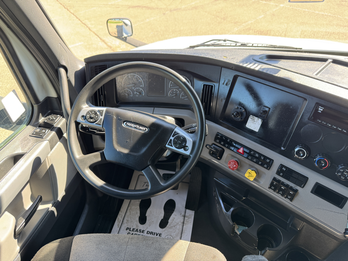 Dashboard and steering wheel of a Freightliner truck cab showing instrument cluster, steering wheel with Freightliner logo, center console controls, and a floor mat that reads 'PLEASE DRIVE CAREFULLY'