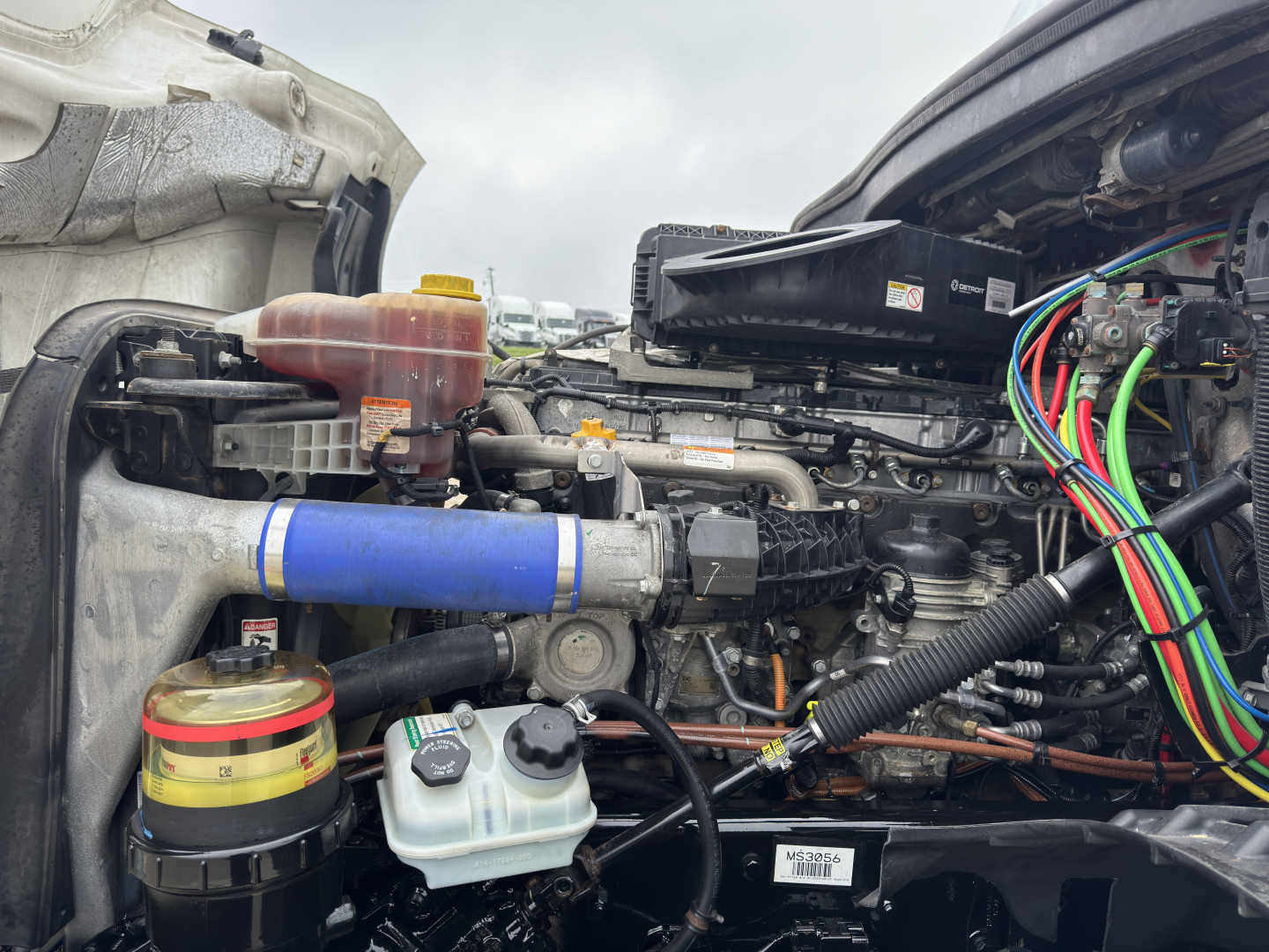 Engine bay of Freightliner Cascadia showing coolant reservoir, blue silicone intercooler hose, various colored wiring harnesses, and fluid reservoirs.