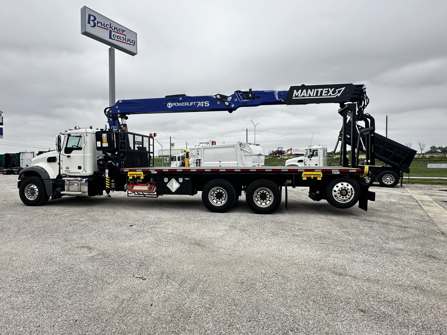 Truck with a blue Manitex hydraulic crane mounted on a white cab and chassis, extended over the trailer bed.