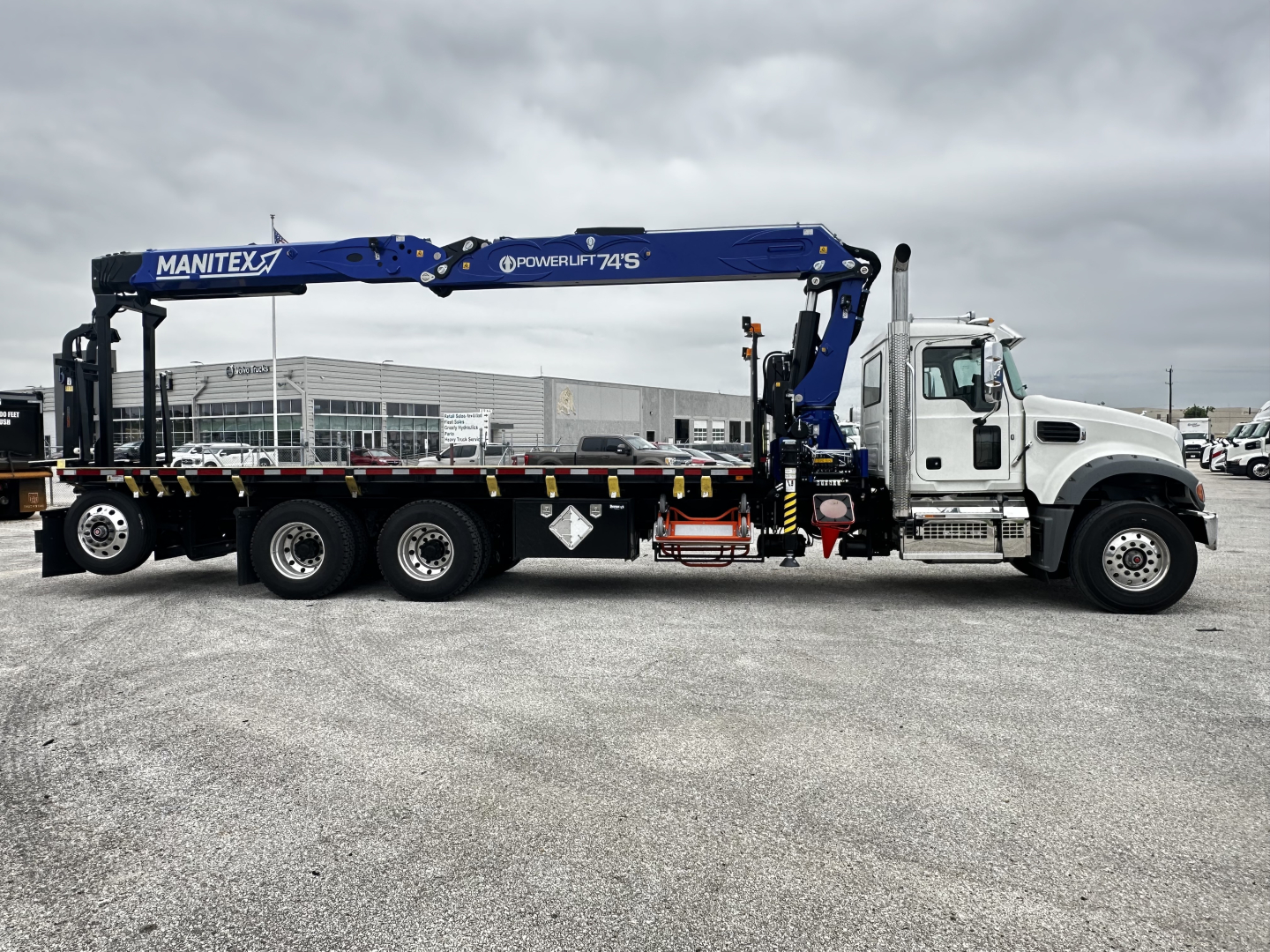 Truck cab and chassis with a blue Manitex PowerLift crane mounted on a white Mack GR64F tractor unit, flatbed and supporting equipment visible.