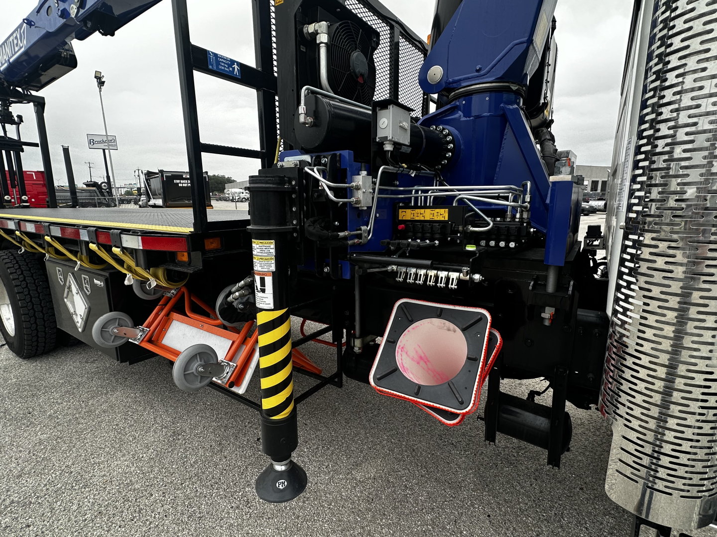 Mack GR64F cab and chassis rear view showing hydraulic stabilizers, controls, and access panels; chrome diamond-plate fuel tank side, warning labels, and landing gear leg.