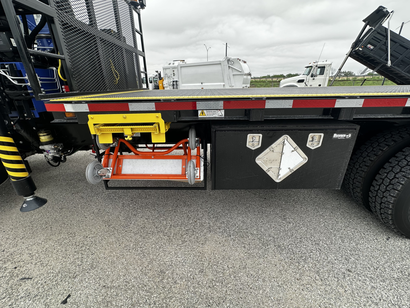 Lightly visible undercab view of a Mack GR64F cab and chassis showing a yellow tool cradle, orange safety reel, and black toolbox with white diamond placard.
