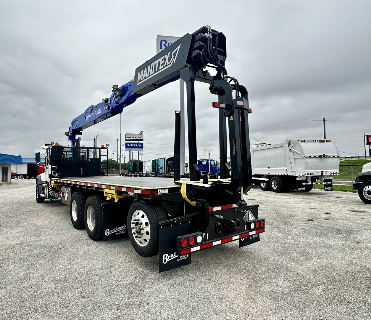 Mack GR64F chassis with Manitex crane mounted on a flatbed trailer, rear view showing stabilizers and tail lights.