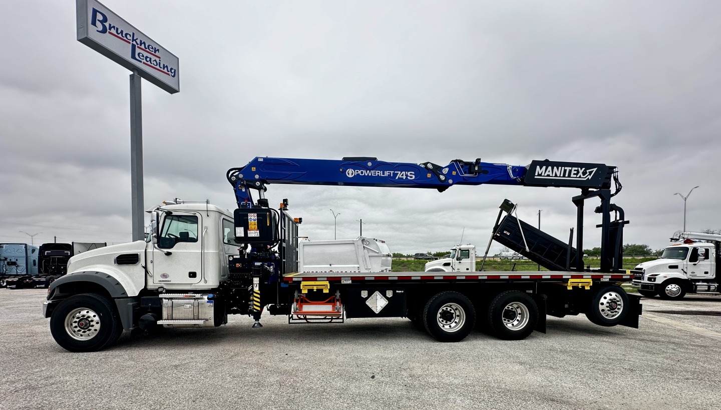Truck cab and chassis with a blue Manitex crane arm extended on a white Mack GR64F with black chassis, white cab, and a flatbed. Visible text includes MANITEX on crane and POWERLIFT on side of arm.