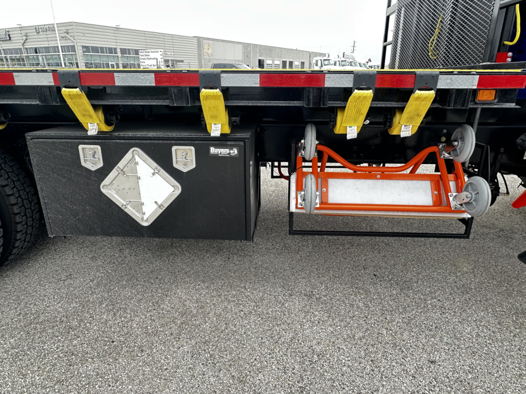 Flatbed truck undercarriage showing fuel/air tanks and side steps with yellow straps and aluminum toolbox on Mack GR64F cab and chassis.