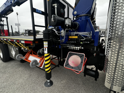 Mack GR64F cab and chassis rear view showing hydraulic stabilizers, controls, and access panels; chrome diamond-plate fuel tank side, warning labels, and landing gear leg.