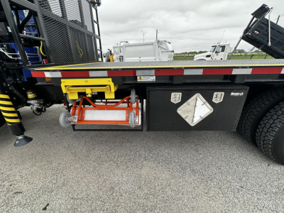Lightly visible undercab view of a Mack GR64F cab and chassis showing a yellow tool cradle, orange safety reel, and black toolbox with white diamond placard.