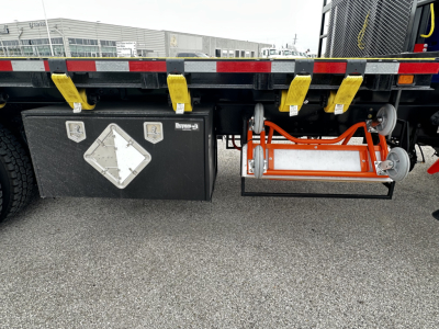 Flatbed truck undercarriage showing fuel/air tanks and side steps with yellow straps and aluminum toolbox on Mack GR64F cab and chassis.