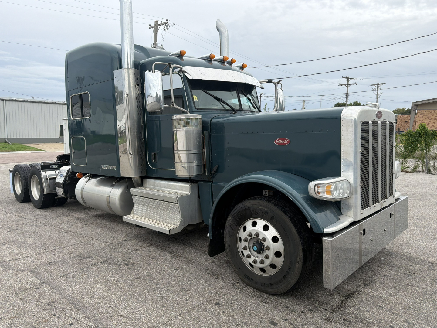 Peterbilt 389 conventional sleeper tractor in blue-gray showing front and side, with chrome exhaust stacks, large dual rear wheels, and silver bumper.