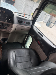 Interior of a 2021 Peterbilt 389 cab showing dashboard with many gauges, control switches, and a black leather driver's seat. Windshield and side mirror visible.