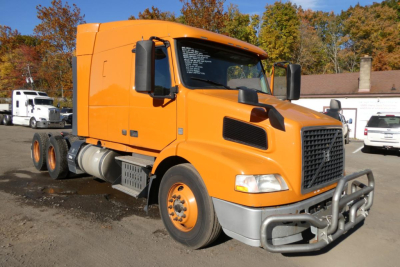 Orange Volvo VNM conventional sleeper truck with black grille guard and large mirrors, side exhaust, in a parking area.