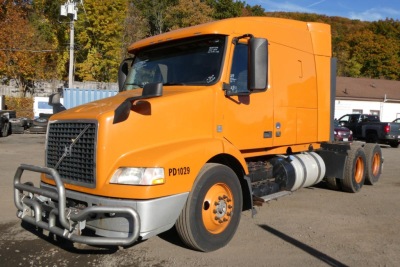 Volvo VNM sleeper tractor in orange, front-left view, showing grille guard, side fuel tanks, and dual rear axles with orange rims.