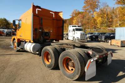 Volvo VNM heavy-duty tractor truck chassis with orange sleeper cab and dual rear axles, dump-style rear cargo area, daytime lot with other vehicles