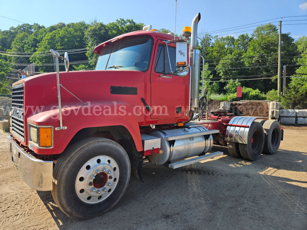 Mack CH613 truck cab in red with front grille and exhaust stacks, side view, showing dual rear axles and fuel tanks; located in a yard with stacked lumber in background.