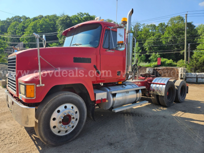 Mack CH613 truck cab in red with front grille and exhaust stacks, side view, showing dual rear axles and fuel tanks; located in a yard with stacked lumber in background.