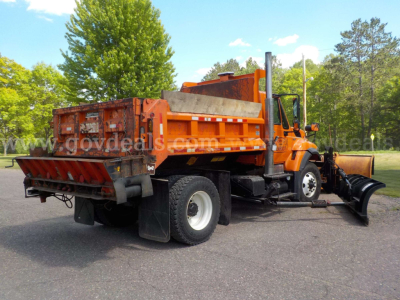 2009 International 7400 plow truck in field, side/back view showing front plow, rear dump bed, orange cab and chassis with black snow plow equipment.