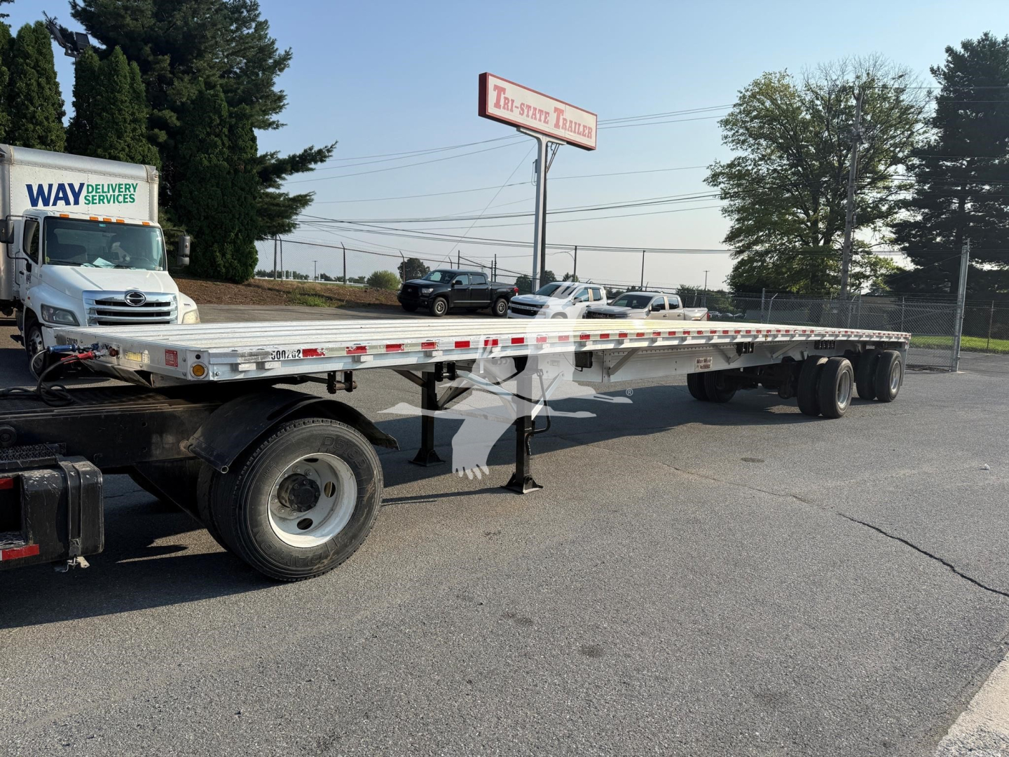 Trailer in a lot showing a long flatbed with dual rear axles and support stands. Visible branding on signage and truck, no text on the trailer readable.