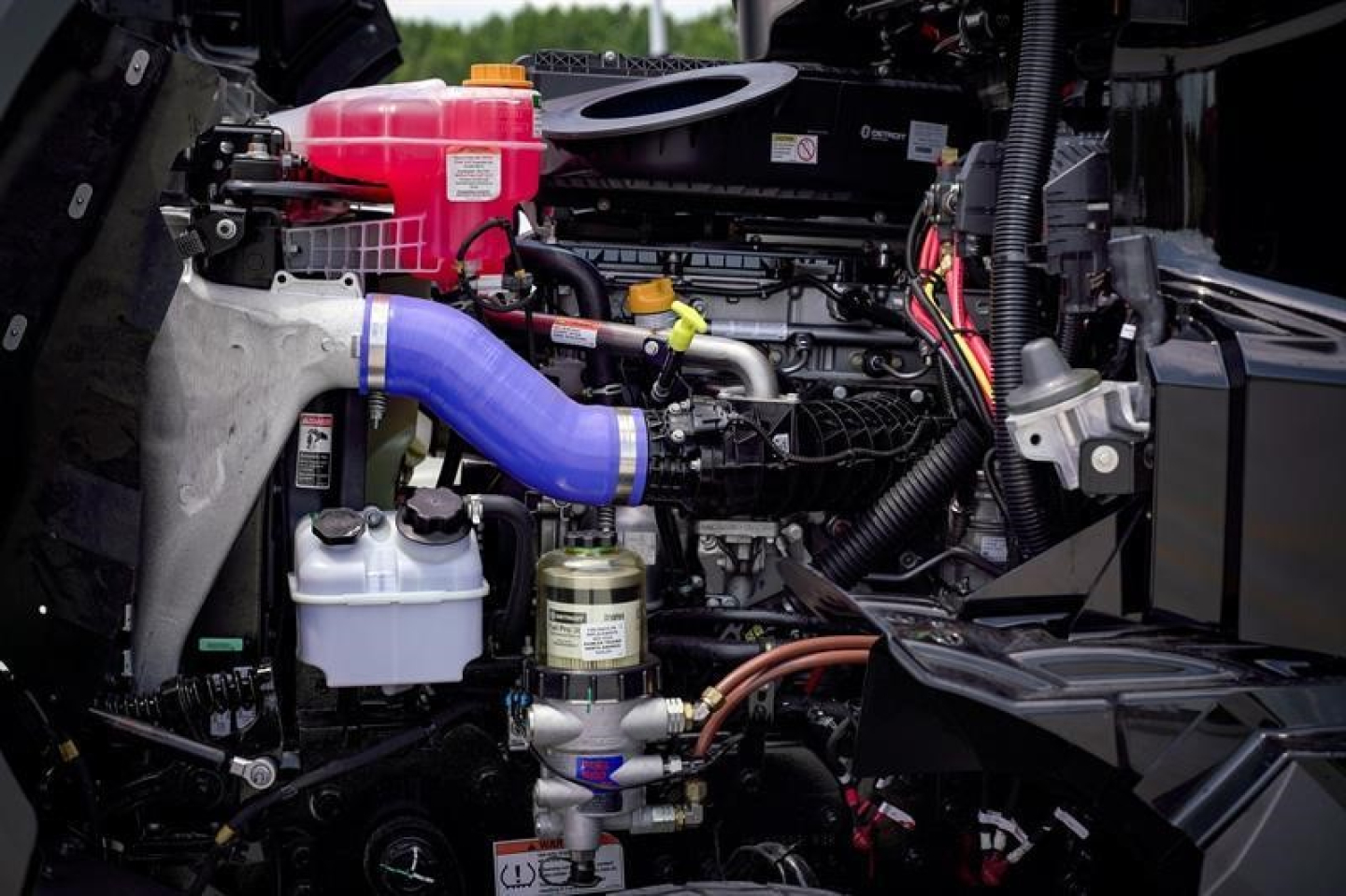 Truck engine bay showing large blue intake hose, pink coolant reservoir, white fluid containers, and various hoses and filters in a Western Star 47X powertrain.