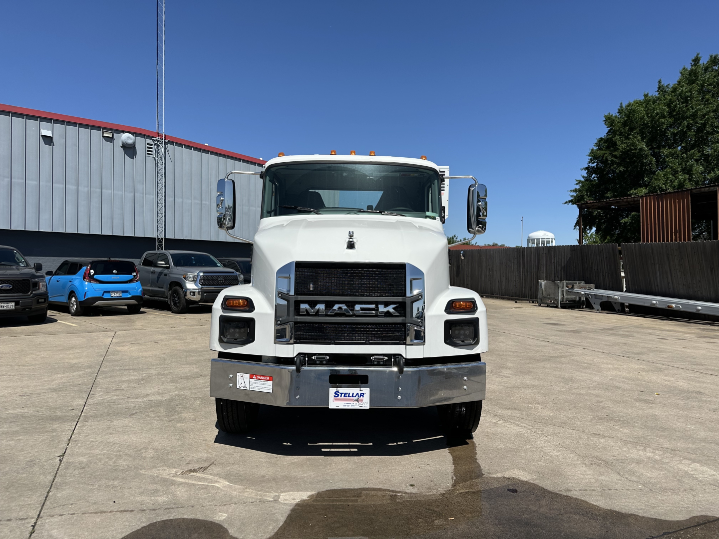 Front view of a white 2026 Mack MD7 truck showing large grille with 'MACK' emblem and bumper; license plate area; service/utility lot in background.