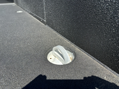 On a Mack MD7 truck bed deck, view of a circular recessed fuel or access cap with a gray twist-lock handle, surrounded by black textured surface.