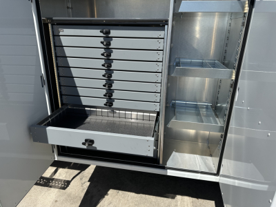 Service body drawers and shelves inside a Mack MD7 utility truck storage compartment showing multiple shallow drawers and vertical tool slots with a pull-out drawer open.