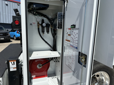 Service/utility truck interior with hydraulic system controls and warning labels visible in an open compartment; includes hoses, gauges, and a red reel inside the bay.