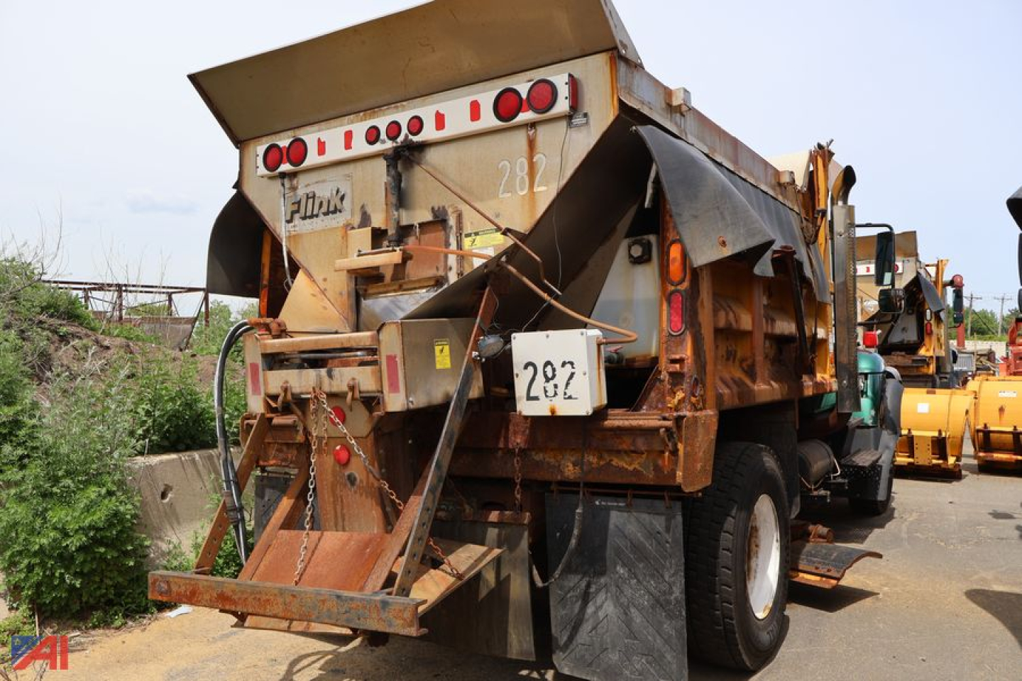 Dump truck from rear angle showing dump bed and taillights, labeled with number plate; rusty metal panels and heavy-duty wheels visible.