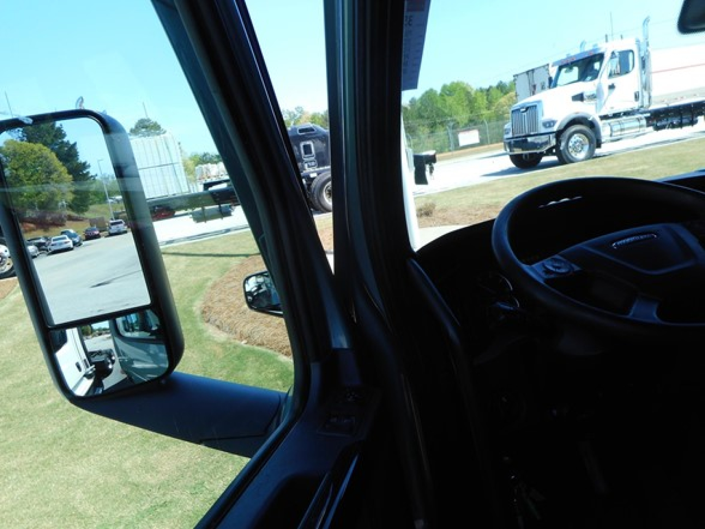 Truck cab interior view of Freightliner with side mirror and windshield, showing truck yard outside.