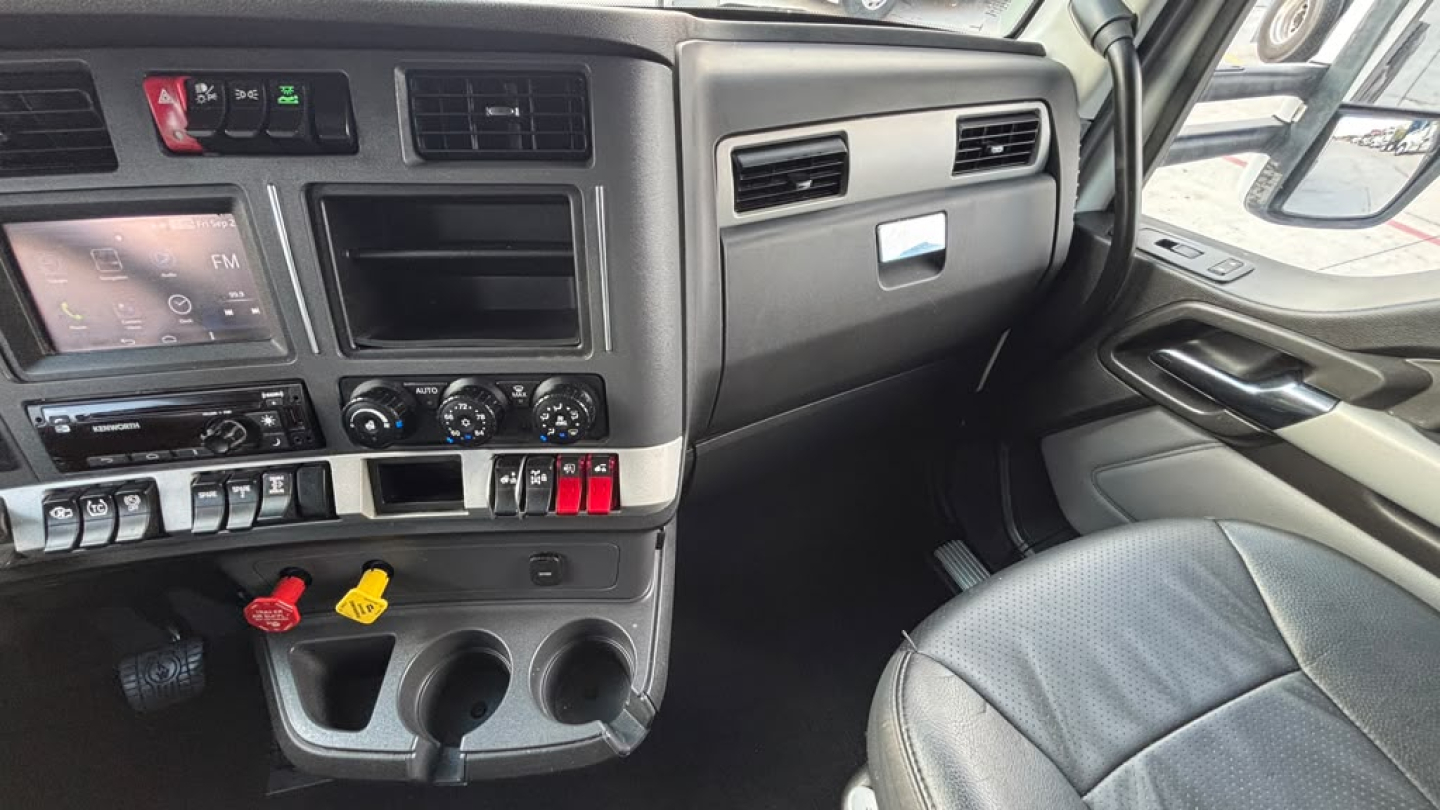 Kenworth truck dash interior showing infotainment screen, climate controls, switches, and cup holders; steering wheel and passenger seat visible.